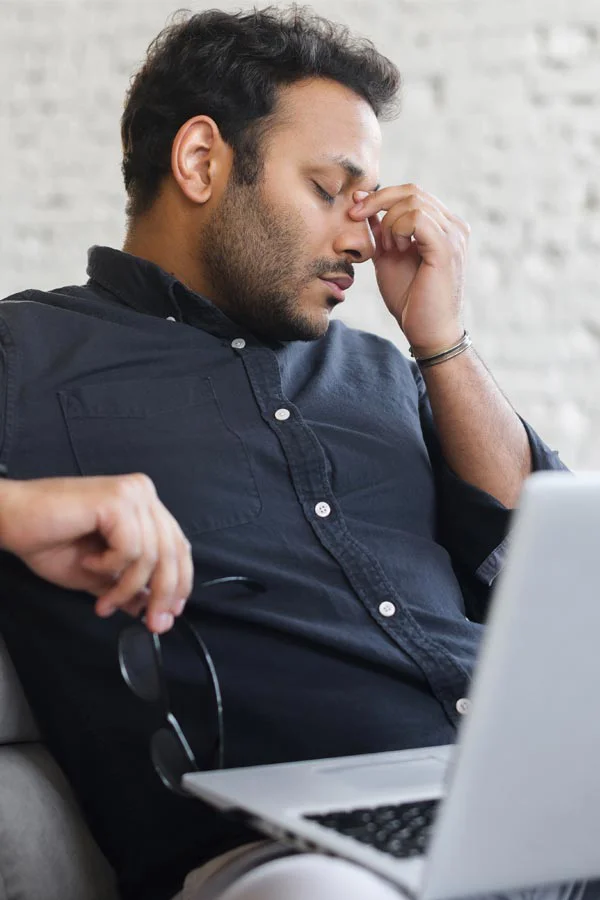 A middle-aged man with a laptop closing his eyes and rubbing his nose. He needs treatment for Adrenal Fatigue Syndrome from Dr. Noelani Rodriguez in San Diego.