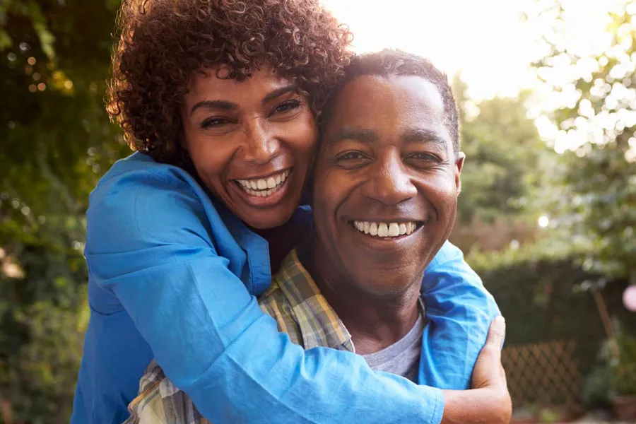 Autoimmune-Doctor A middle-aged African American couple outside, the woman embracing the man from behind pressing her cheek to his, both are smiling due to successful Autoimmune Disorder treatment from Dr. Noelani Rodriguez in San Diego.