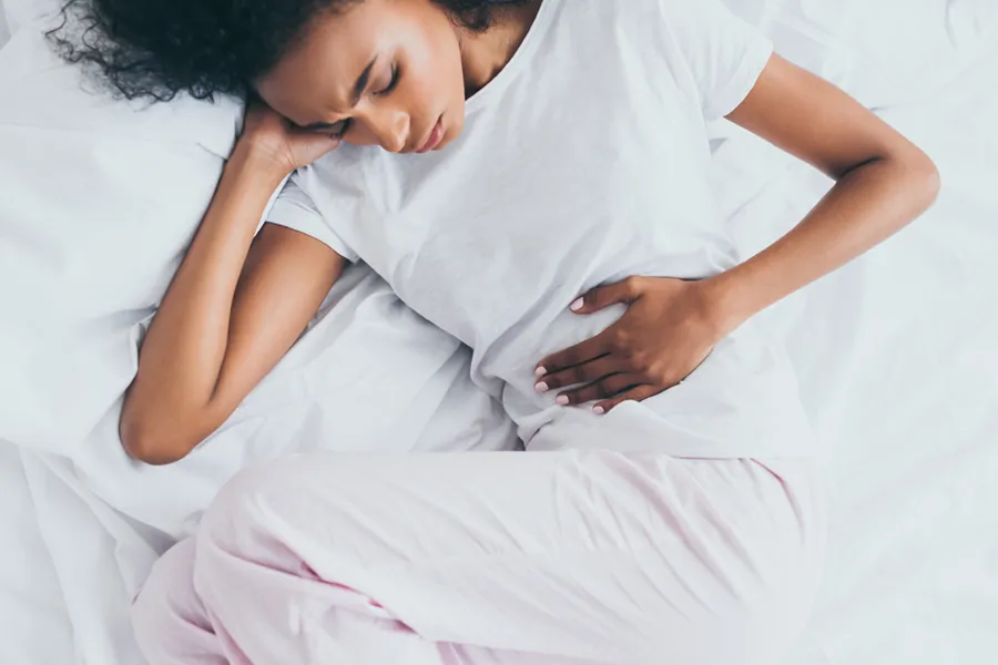Crohns-Disease-Clinic A dark-skinned woman in white clothes lays on her bed clutching her middle before getting treatment for Crohn's Disease from Dr. Noelani Rodriguez in San Diego.