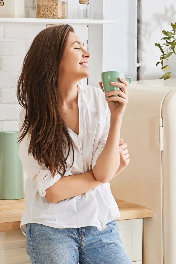 Crohns-Disease-Treatment A woman with a white blouse an jeans drinking tea in her kitchen, smiling after getting treatment for Crohn's Disease from Dr. Noelani Rodriguez in San Diego.