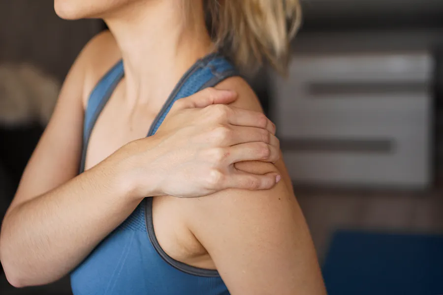 Frozen-Shoulder-Clinic A close-up view of a woman in a blue tank top, gripping her own shoulder with a look of discomfort. Receive expert Frozen Shoulder treatment by Dr. Noelani Rodriguez located in San Diego.