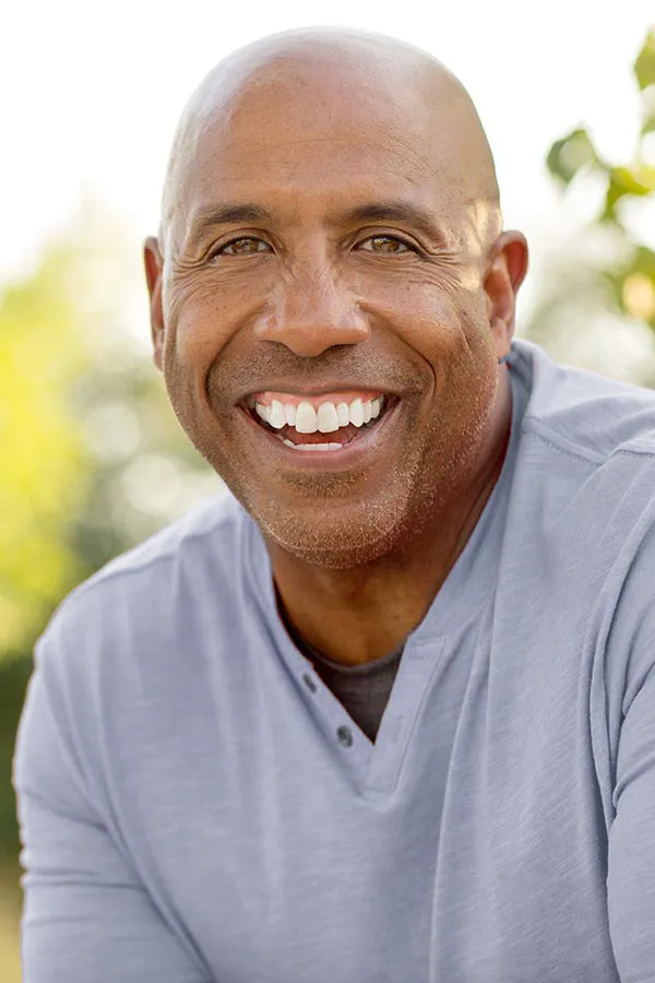 Healthy-Aging-Peptides-Treatment A middle-aged black man in a gray v-neck t-shirt sits outside smiling, happy with the healthy aging peptide treatments from Dr. Noelani Rodriguez in San Diego