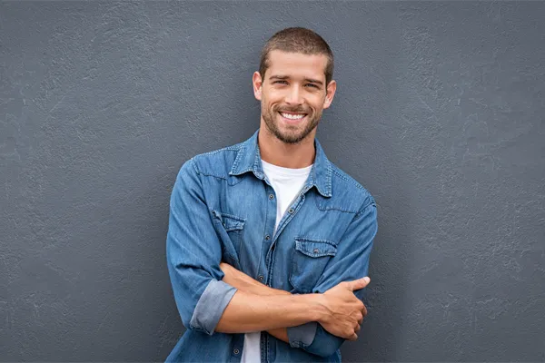 Hormones-for-Men-Doctor A man in a denim shirt stands smiling against a gray-blue wall, pleased with his testosterone hormone therapy from Dr. Noelani Rodriguez in San Diego.