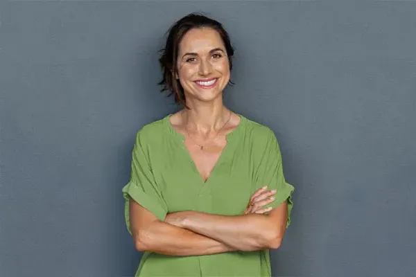 Hormones-for-Women-Doctor A woman in a green shirt stands smiling against a gray-blue wall, pleased with her hormone therapy from Dr. Noelani Rodriguez in San Diego.