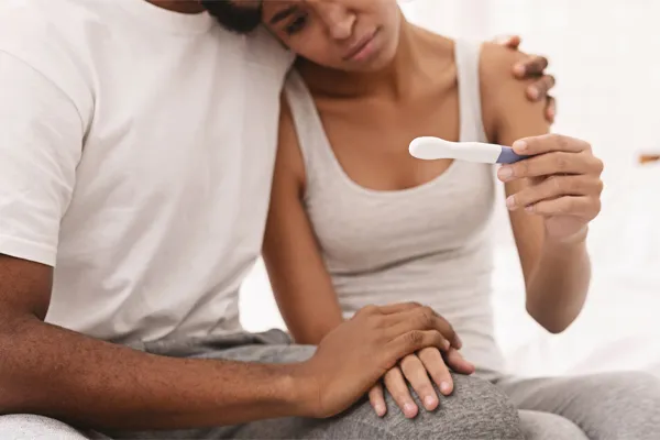Infertility-Clinic A black couple sits on a bed, disappointed with the negative results of a pregnancy test. Get treatment for infertility from Dr. Noelani Rodriguez in San Diego.