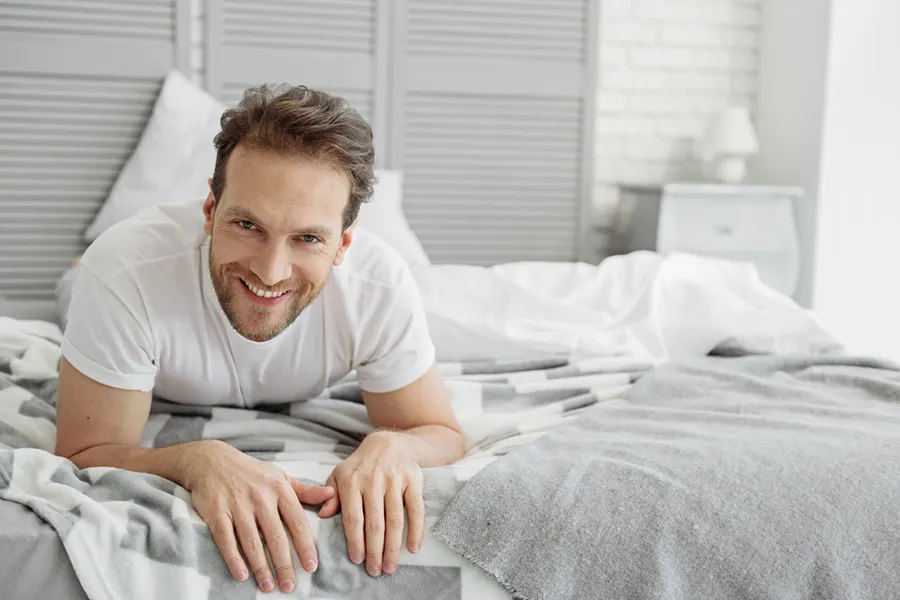 Low-Libido-in-Men-Doctor A dirty-blonde haired man lays on his stomach in bed smiling. Schedule treatment for low libido in men from Dr. Noelani Rodriguez in San Diego.
