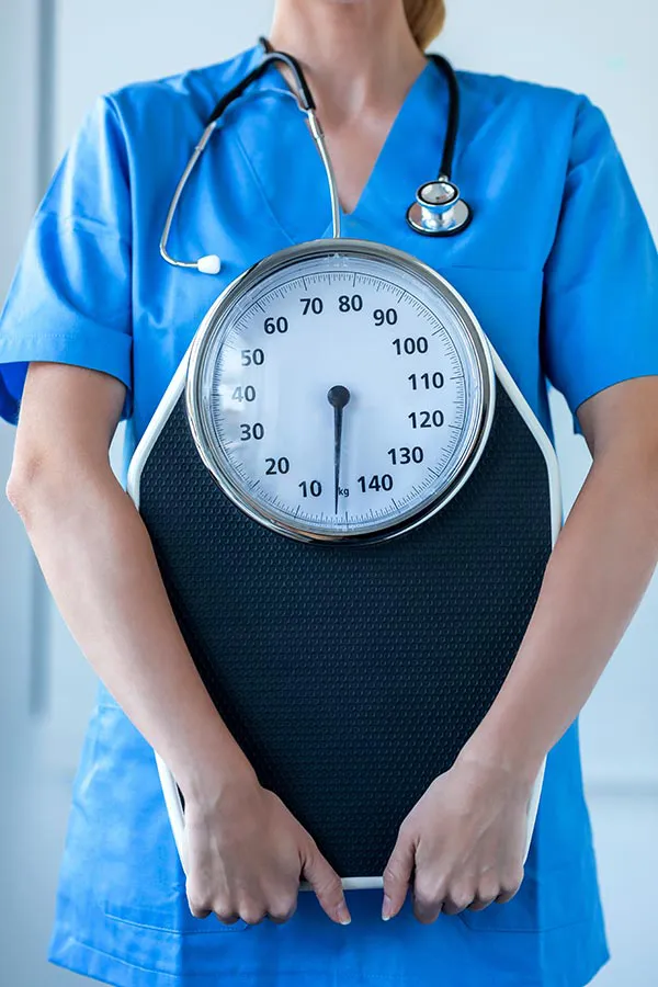 Medical-Weight-Loss-Treatment A nurse in blue scrubs with a stethoscope hanging around her neck holds a scale, representing weight loss treatments from Dr. Noelani Rodriguez in San Diego.