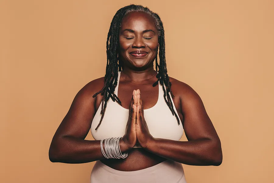 Obesity-Clinic An older African American woman confidently posing against a warm background smiling with hands in a prayer pose. Dr. Noelani Rodriguez treats obesity using functional medicine in San Diego.
