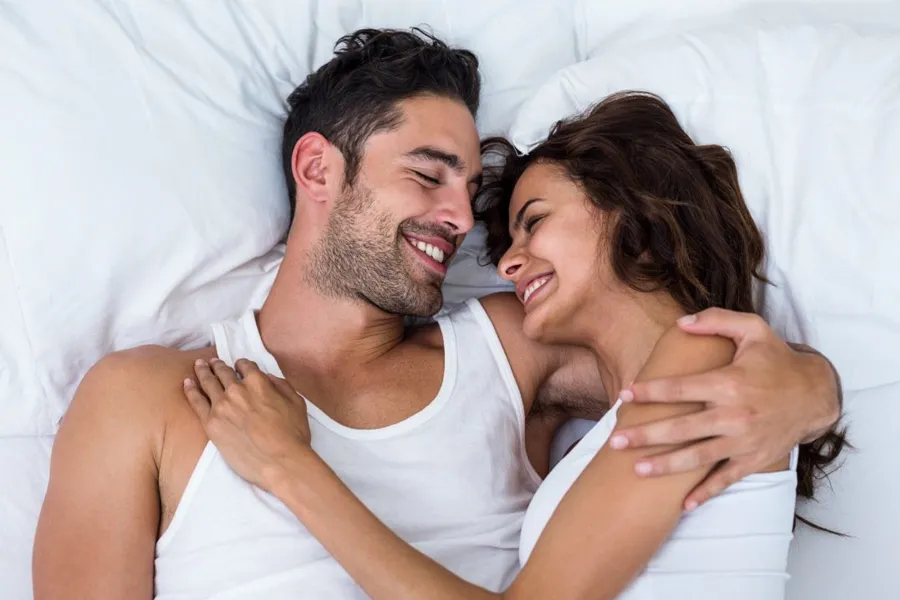 PT141-for-Men-Doctor A laughing man and woman in undershirts lie in each other's arms on a white bed, representing successful PT141 sexual wellness therapy for men from Dr. Noelani Rodriguez in San Diego.