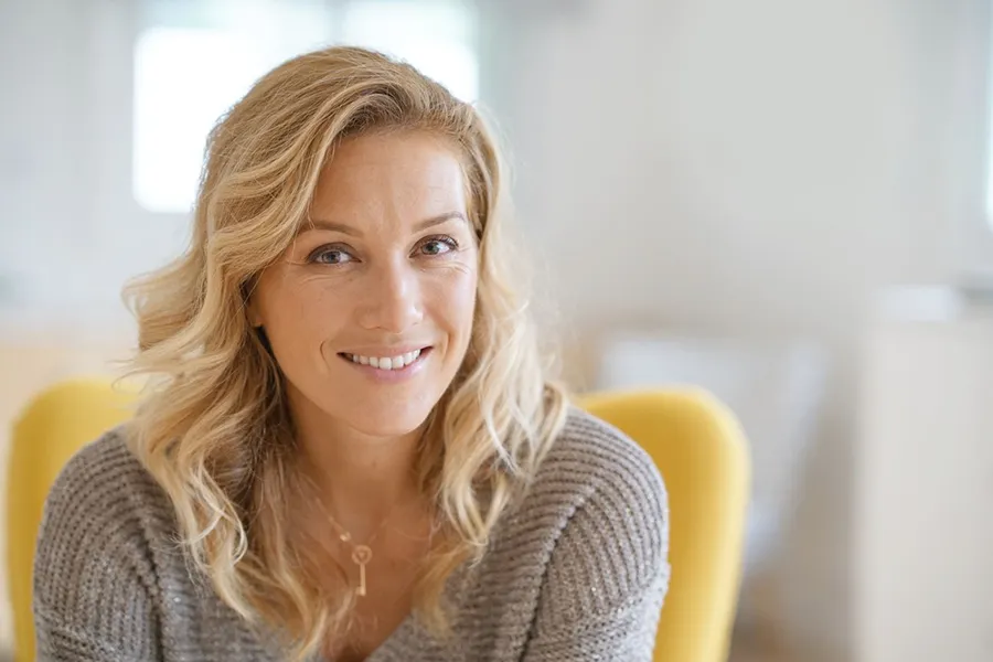 Perimenopause-Doctor A blonde woman wearing a a gray sweater sits inside in a yellow chair, smiling and happy with her perimenopause treatment from Dr. Noelani Rodriguez in San Diego.
