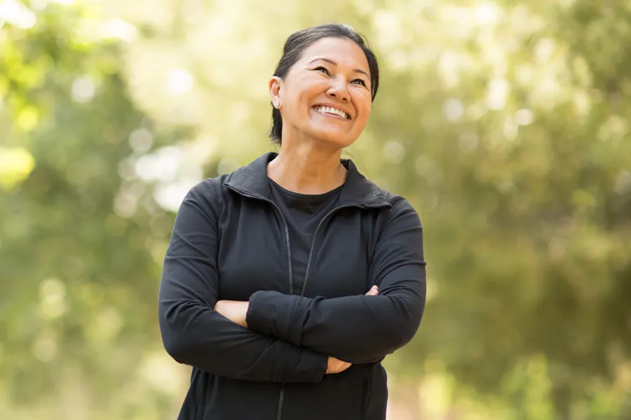 Sermorelin-Ipamorelin-Doctor A middle-aged asian woman in a black warm up stands outside among the trees with her arms folded, smiling and happy with her Sermorelin and Ipamorelin treatments from Dr. Noelani Rodriguez in San Diego.