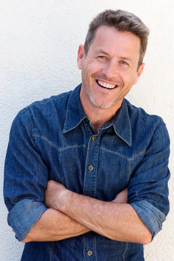 Sermorelin-Ipamorelin-Treatment A middle-aged caucasian man in a denim shirt stands against a white wall with his arms folded, smiling and happy with his Sermorelin and Ipamorelin treatments from Dr. Noelani Rodriguez in San Diego