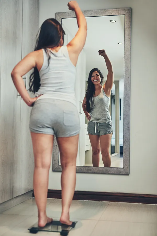 Tirzepatide-Treatment A woman in workout clothes stands on a scale looking in the mirror and pumping her hands in celebration of her successful Tirzepatide treatment from Dr. Noelani Rodriguez in San Diego.
